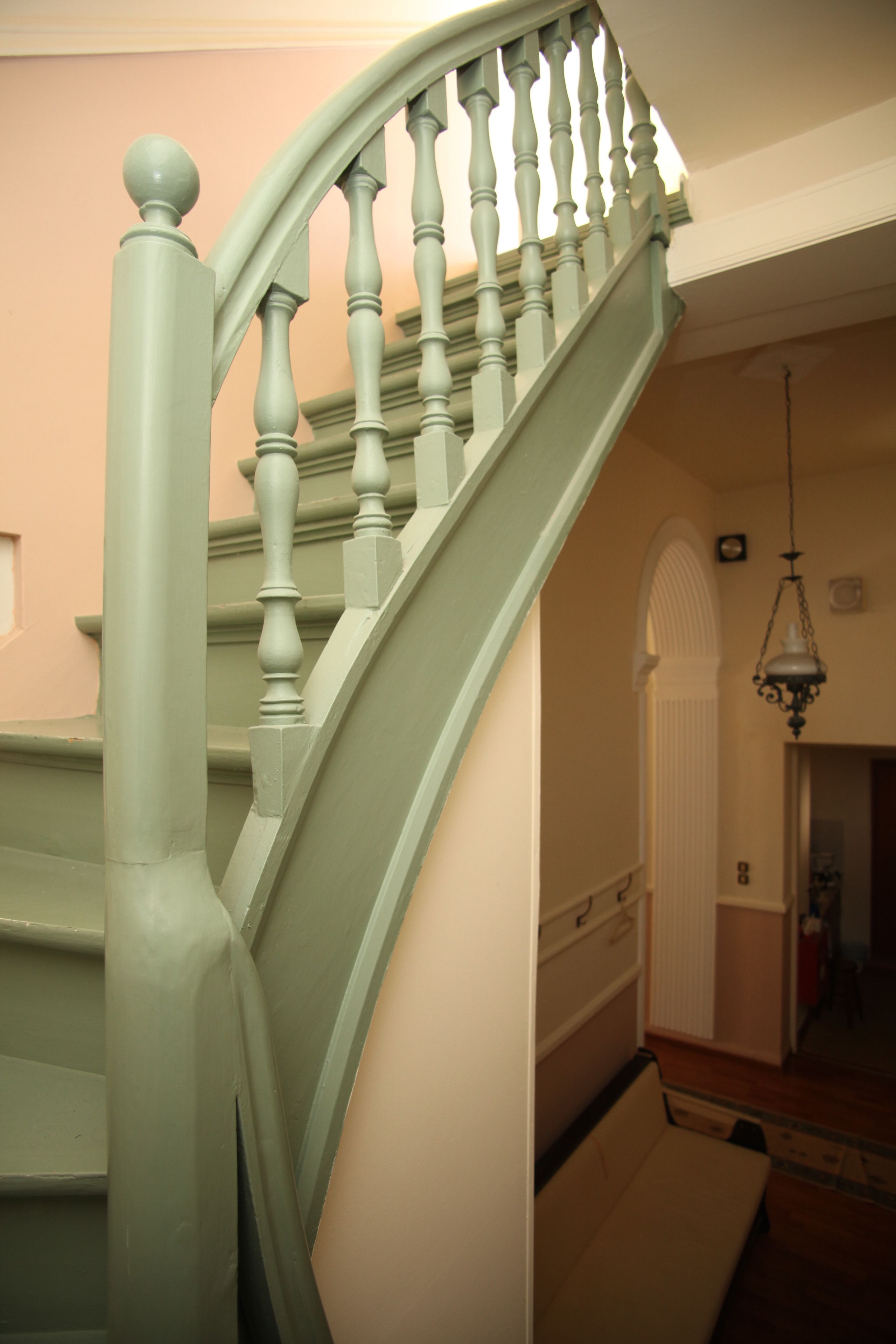 A staircase with a light green handrail and balusters, leading to a landing with a white door and a hanging light fixture.