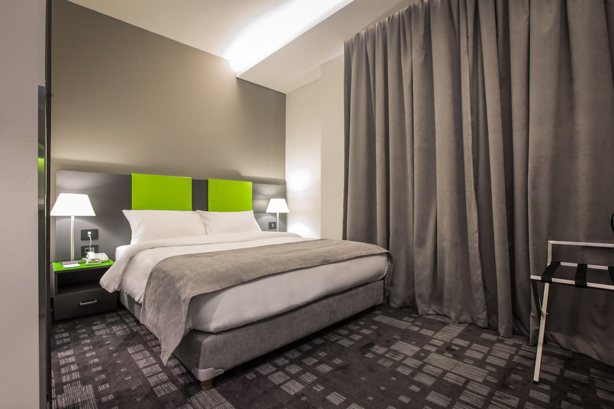 A hotel room with a bed, two bedside tables with lamps, a grey curtain, and a small table with a chair.