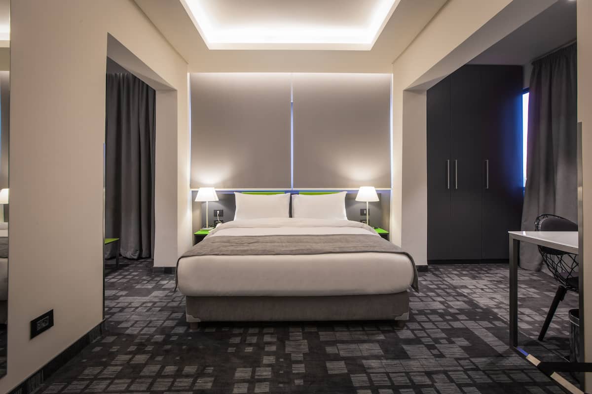 A modern hotel room with a large bed, two bedside tables with lamps, a desk, and a wardrobe.
