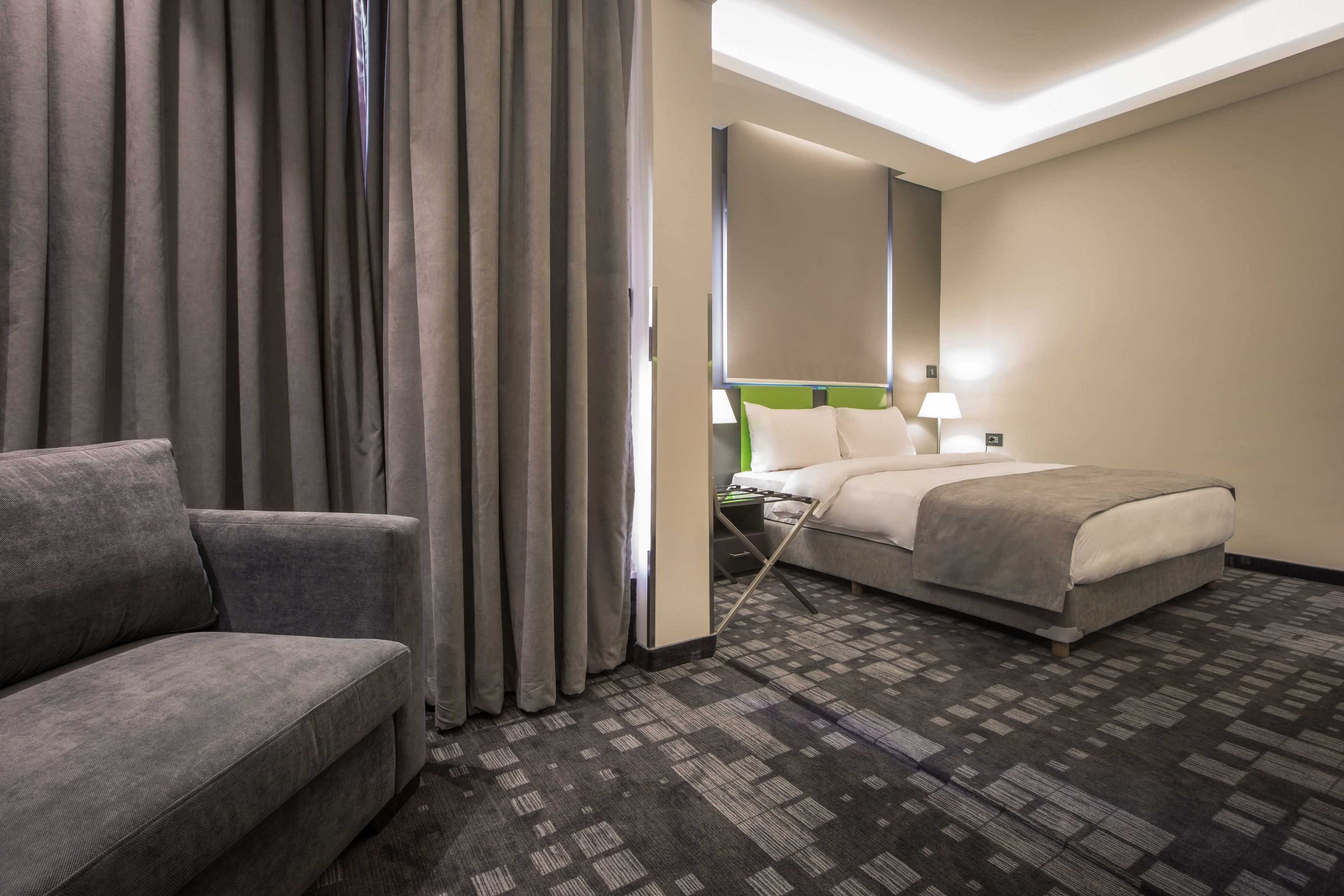 Una habitación de hotel con un sofá gris, una cama con cabecera verde y una alfombra estampada.