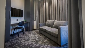 A modern hotel room with a grey sofa, a black chair, a desk, and a flat-screen TV mounted on the wall.