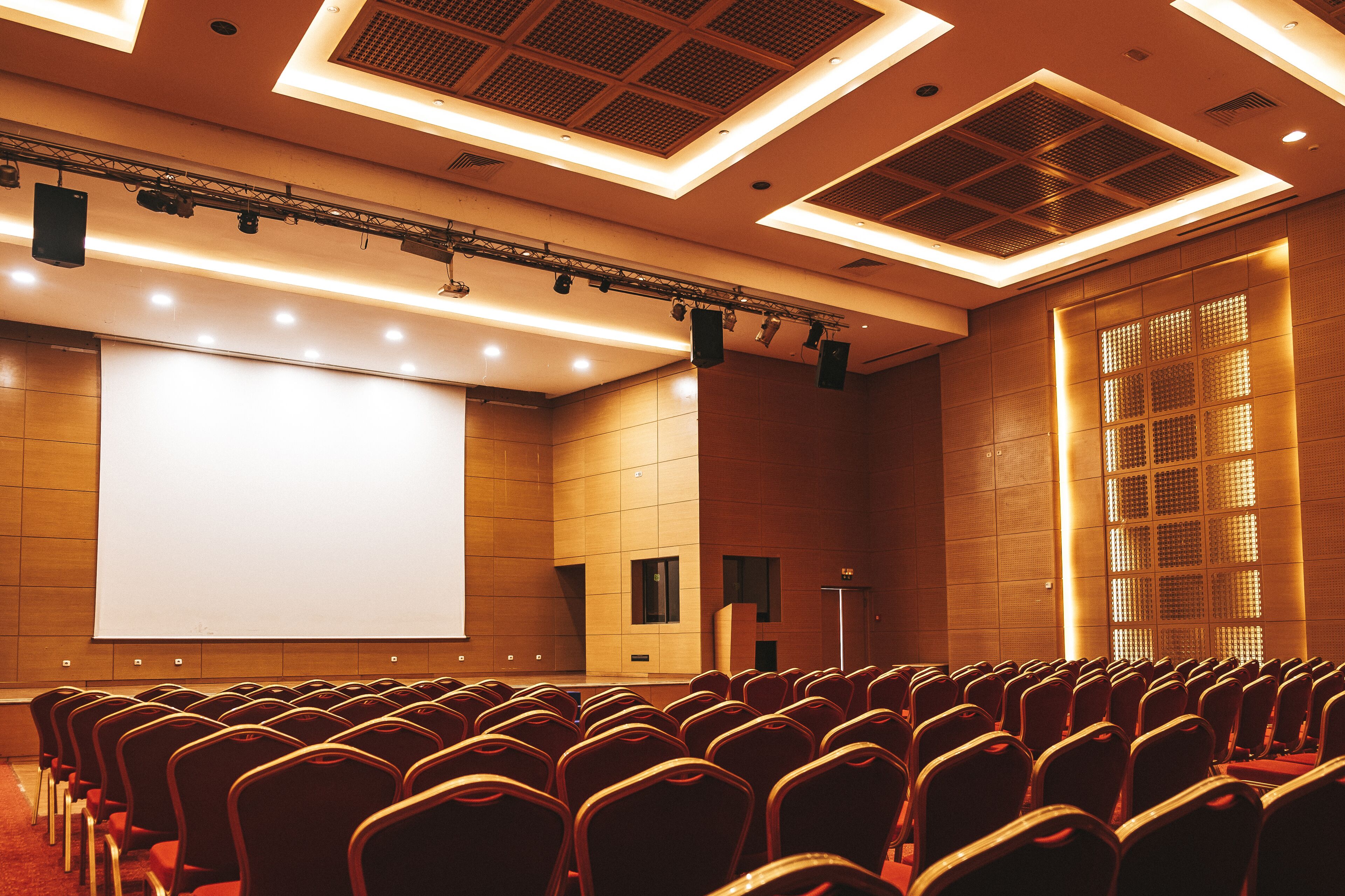 Sala de conferências com fileiras de cadeiras voltadas para uma tela de projeção, equipada com iluminação no teto e sistema de som.