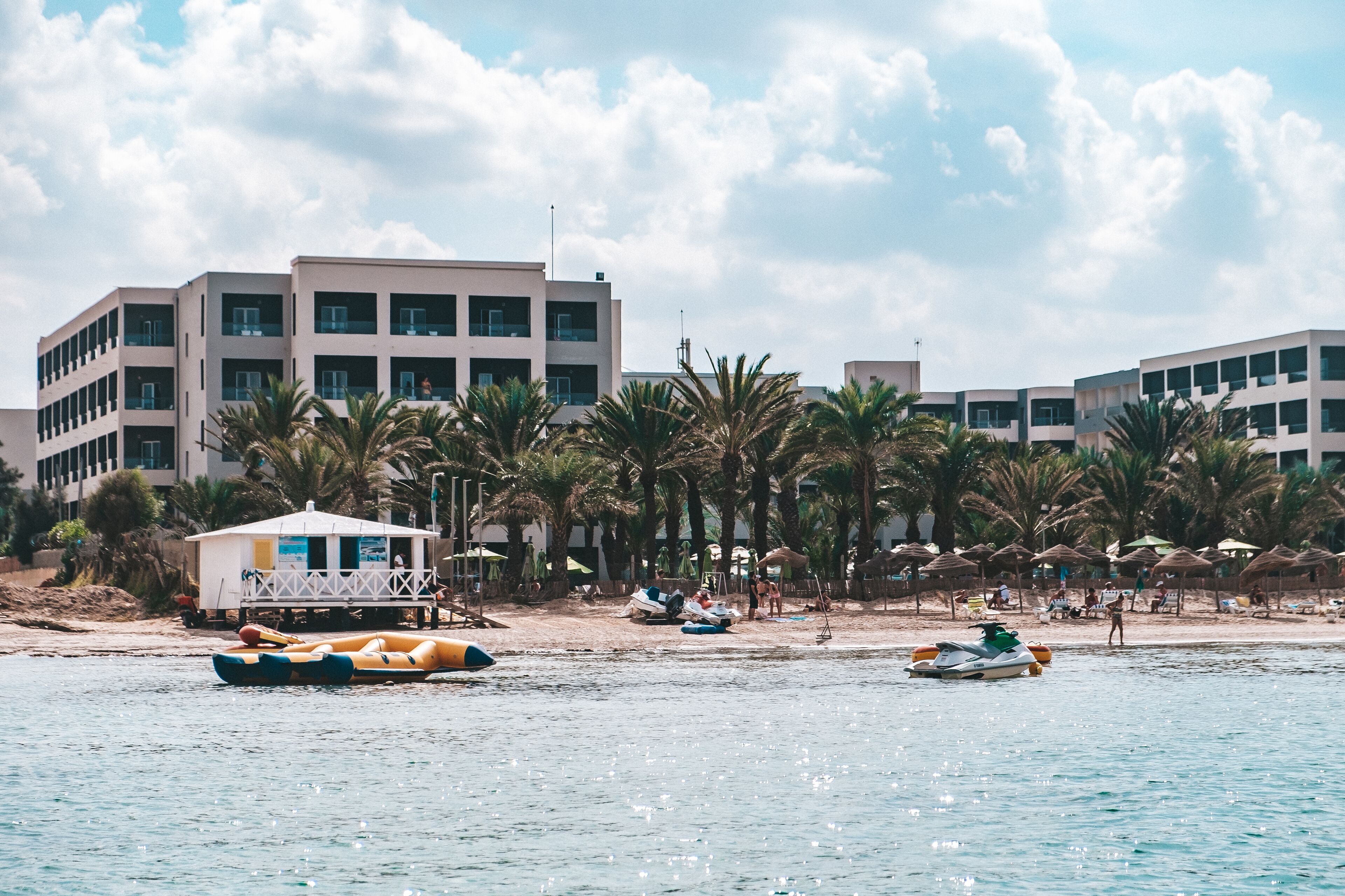 A coastal hotel with multiple buildings, palm trees, and beach umbrellas.