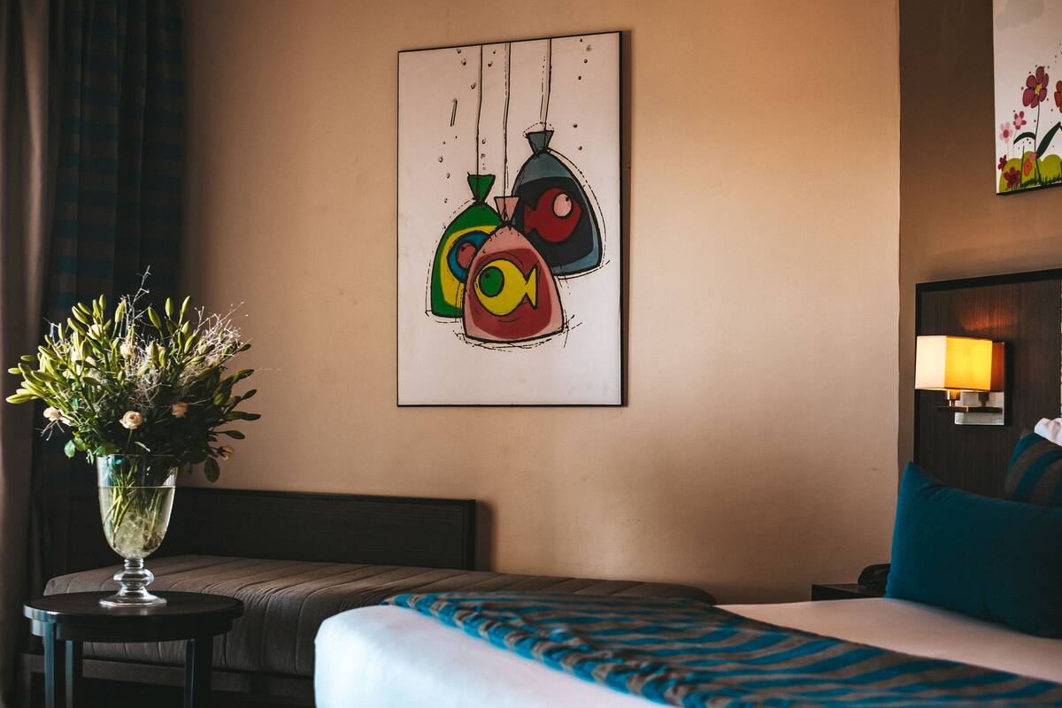 A hotel room with a bed, a bedside table with a vase of flowers, a wall-mounted painting, and a nightstand with a lamp.