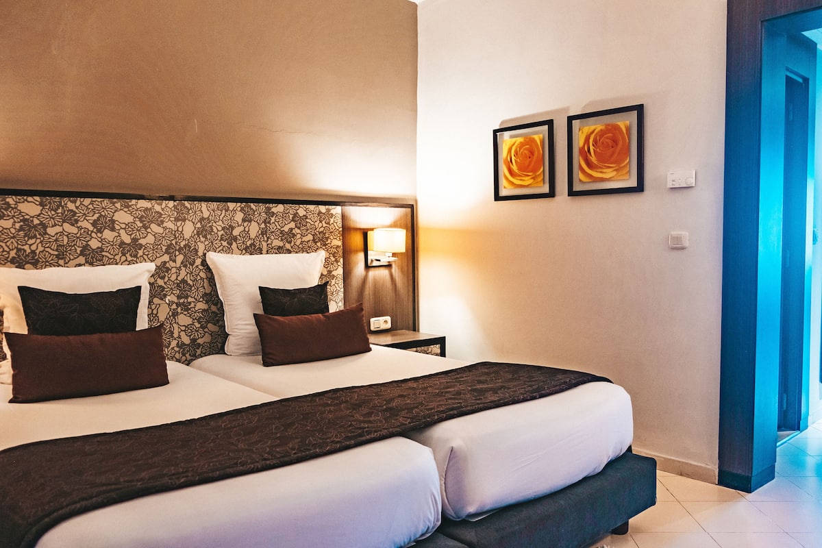 A double bed with a dark headboard, white linens, and brown pillows. There are two wall-mounted lamps and two framed pictures on the wall.