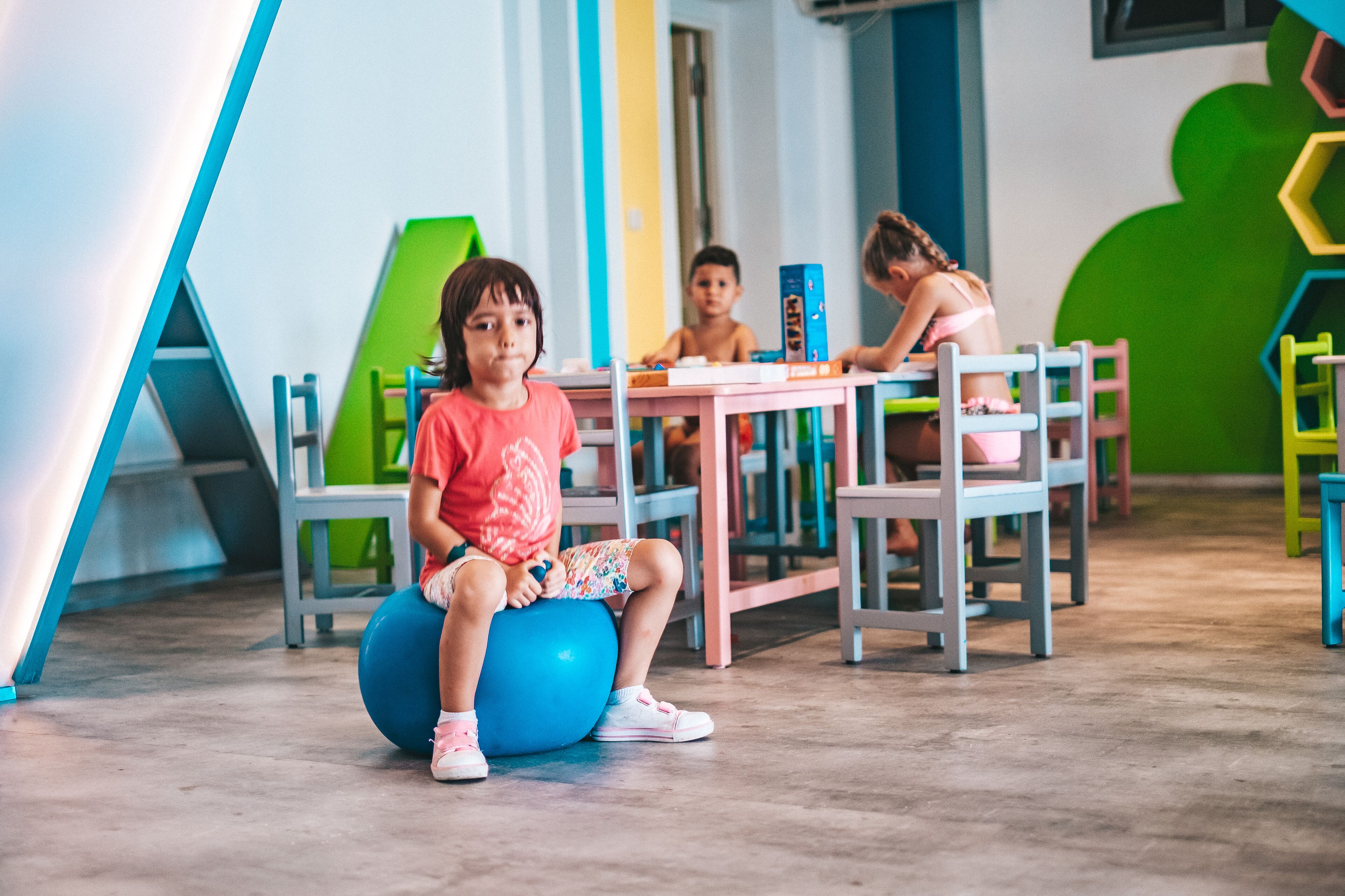 Un niño sentado en una pelota de ejercicios azul en una habitación de colores vivos con una mesa y sillas.