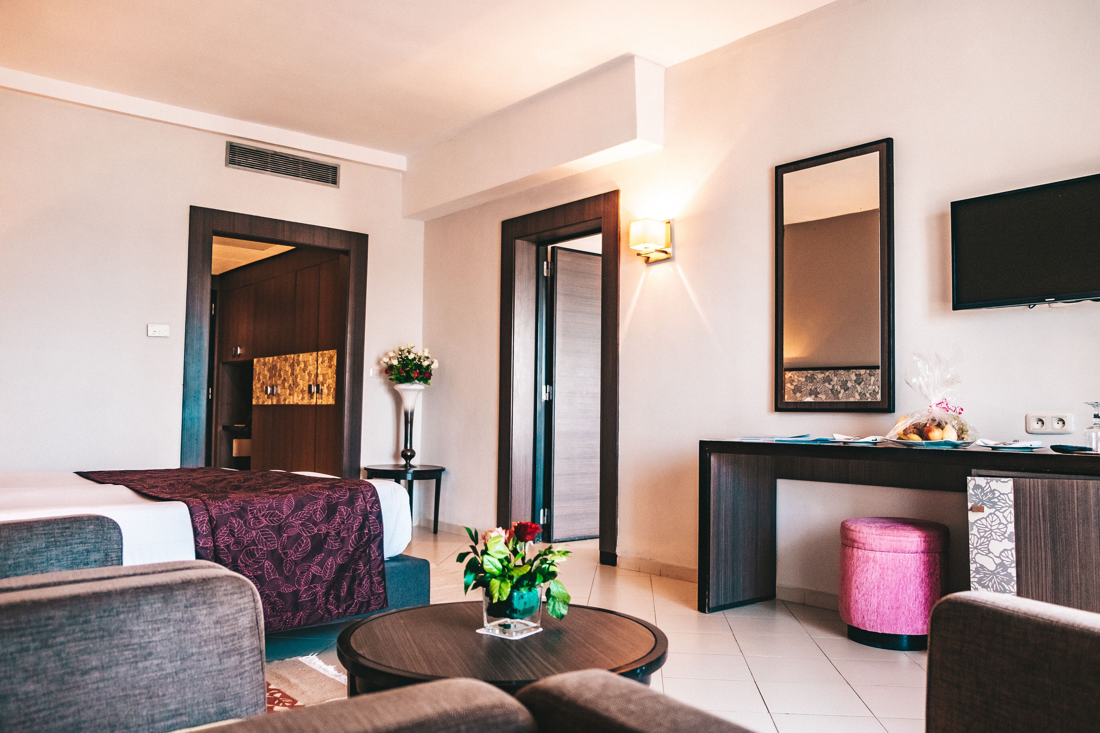 A hotel room with a bed, a desk, a chair, a round table, and a small pink stool.