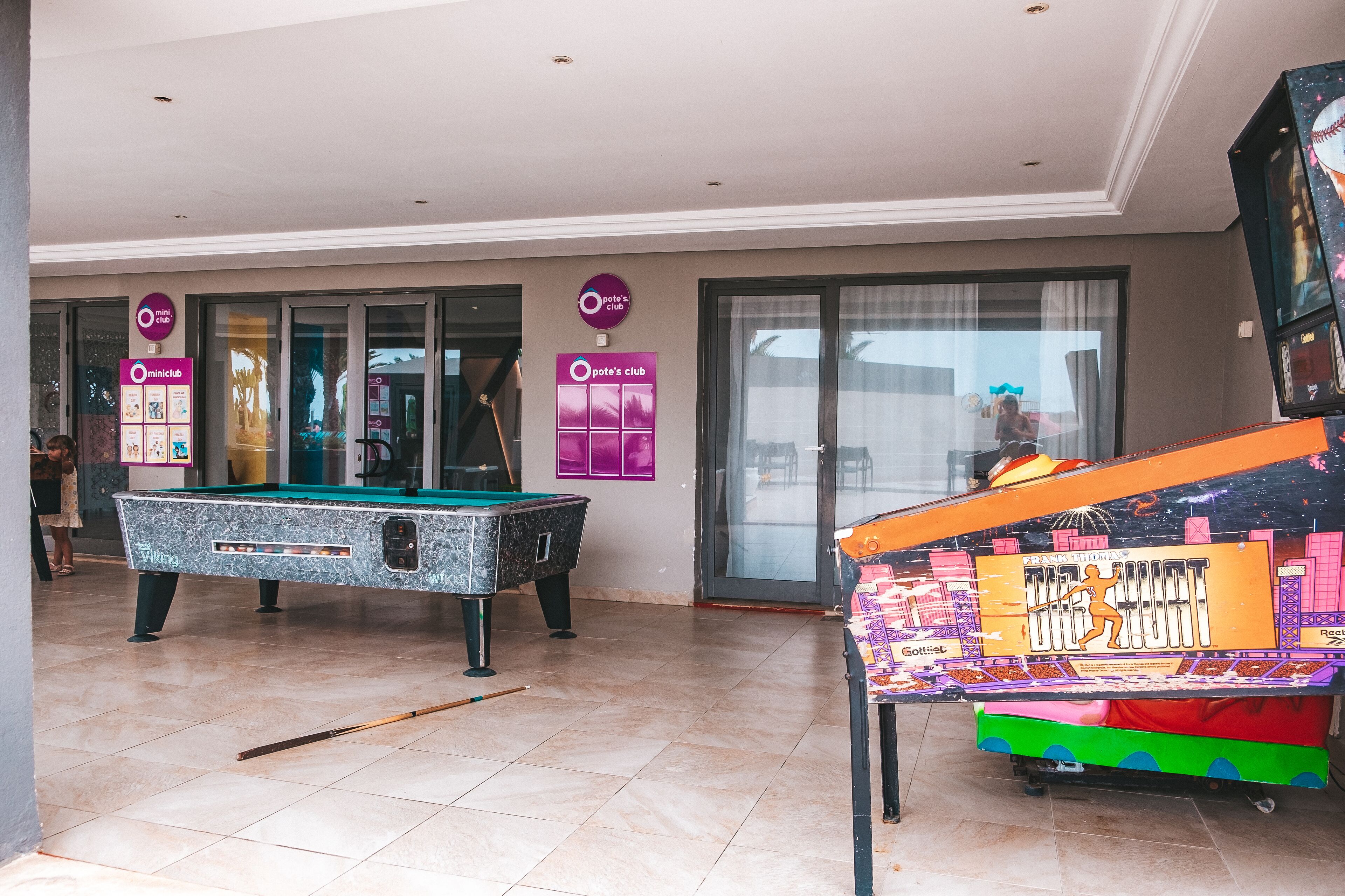 Una mesa de billar, una mesa de pool y un dardo en una sala con ventanas amplias y un letrero que dice 'Pote's Club'.