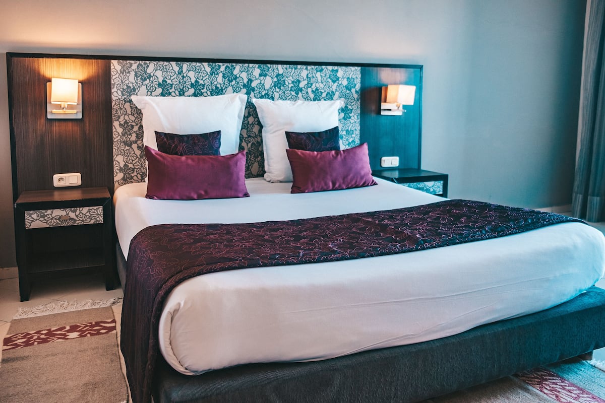 A hotel room with a large bed, two bedside tables, a nightstand, a patterned headboard, and wall-mounted lamps.