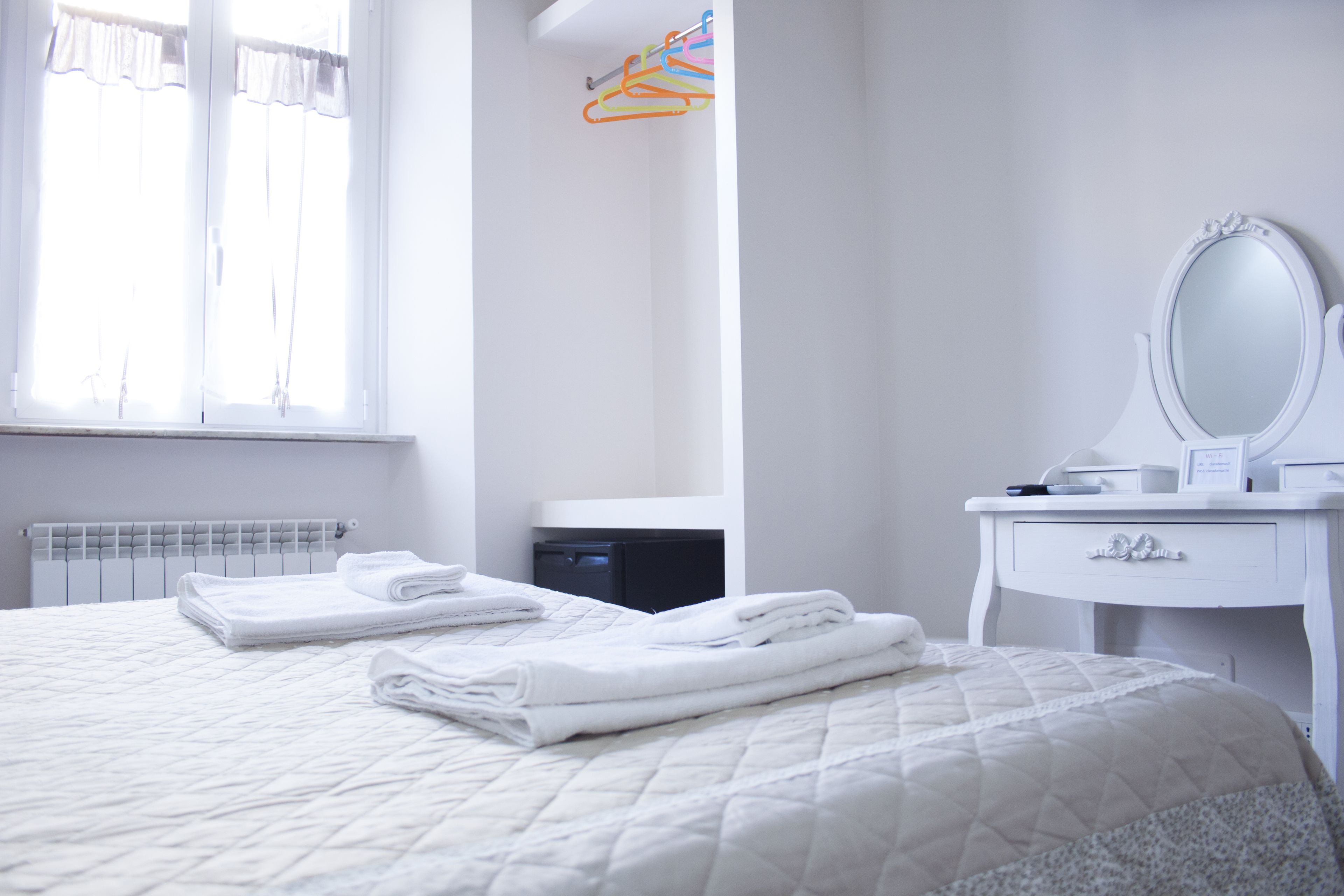 A neatly made bed with folded white towels, a white dresser with a mirror, and a radiator below a window with sheer curtains.