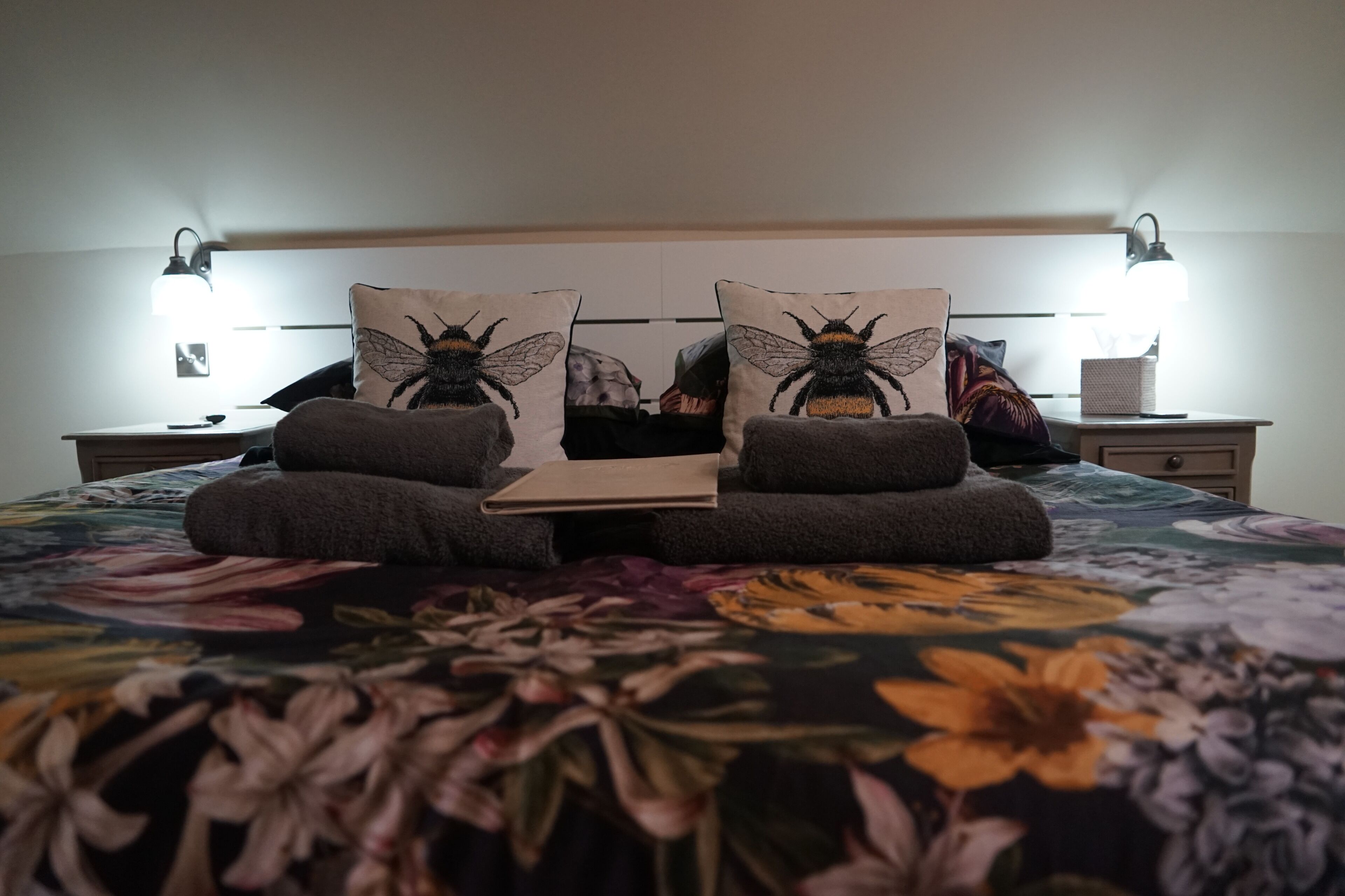Ein Bett mit blumigem Überwurf, zwei Kissen mit Bienenmotiv, ein Nachttisch mit Lampe und ein Beistelltisch ebenfalls mit Lampe.