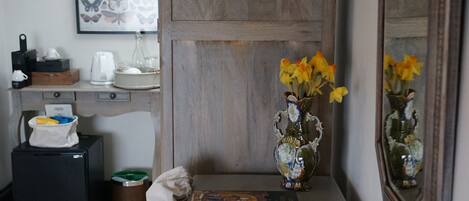 Una habitación con puerta de madera, un escritorio con un jarrón de flores, un cuadro en la pared y una mesita con un libro.