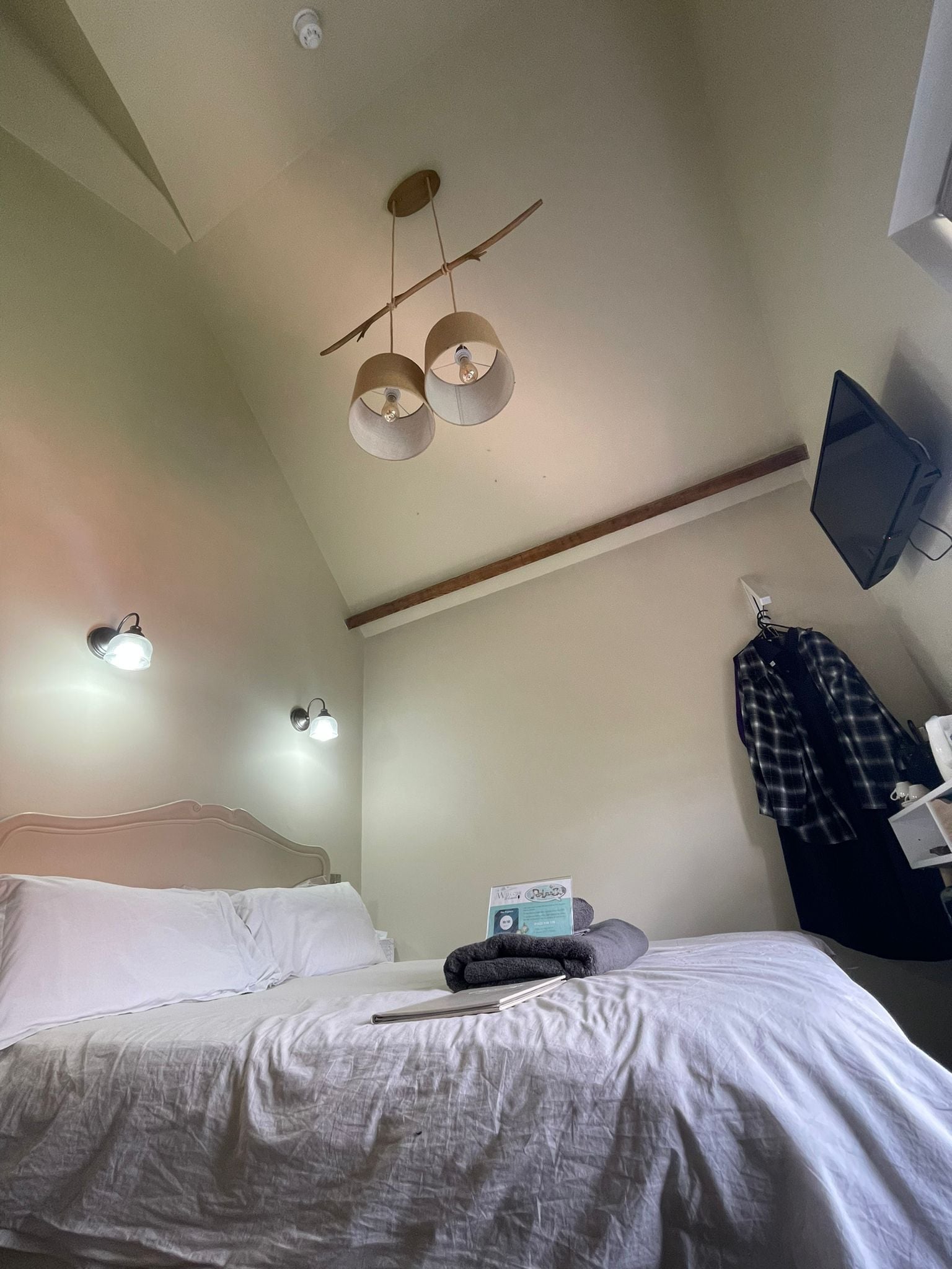 A bedroom with a bed, a desk, a chair, a lamp, a clock, and a TV mounted on the wall.