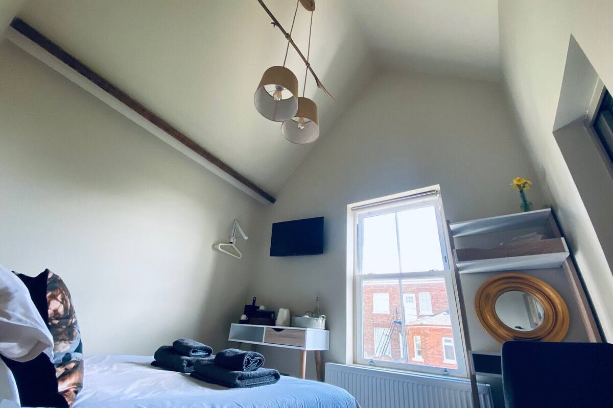 A bedroom with a bed, a window, a radiator, a shelf with a mirror, and a hanging light fixture.