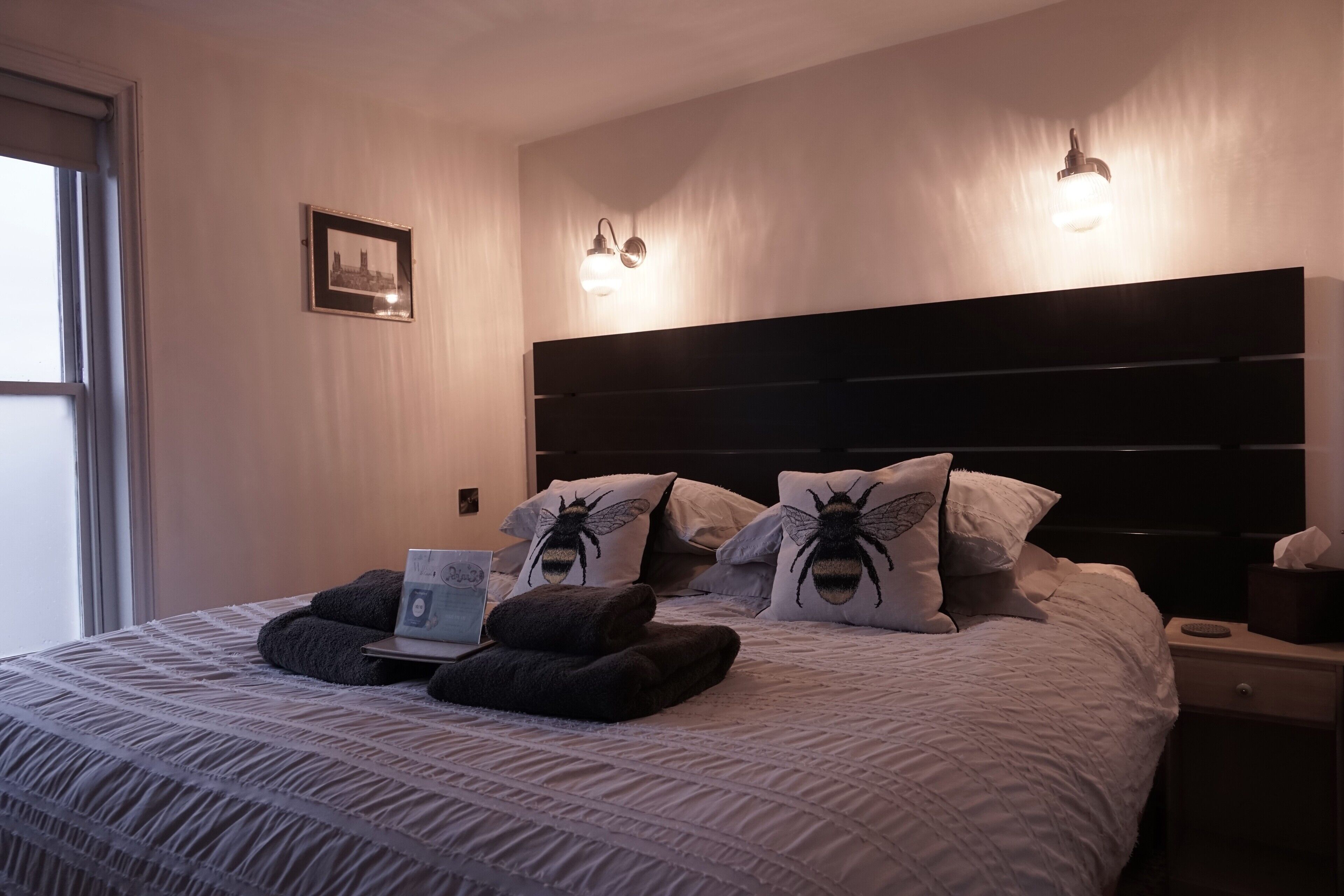 A neatly made bed with a laptop, two pillows with a bee design, and a bedside table with a tissue box.