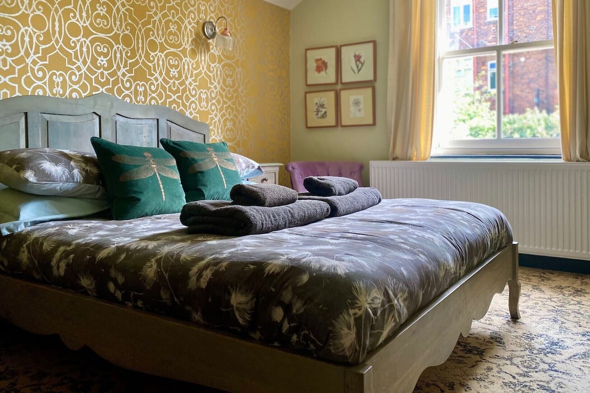 A bedroom with a patterned wallpaper, a bed with floral bedding, and a window with a view of buildings.