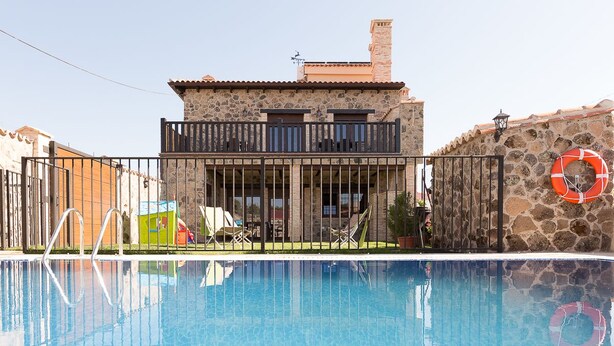 A swimming pool with a stone building in the background, a lifebuoy on the wall, and a fenced area with lounge chairs.