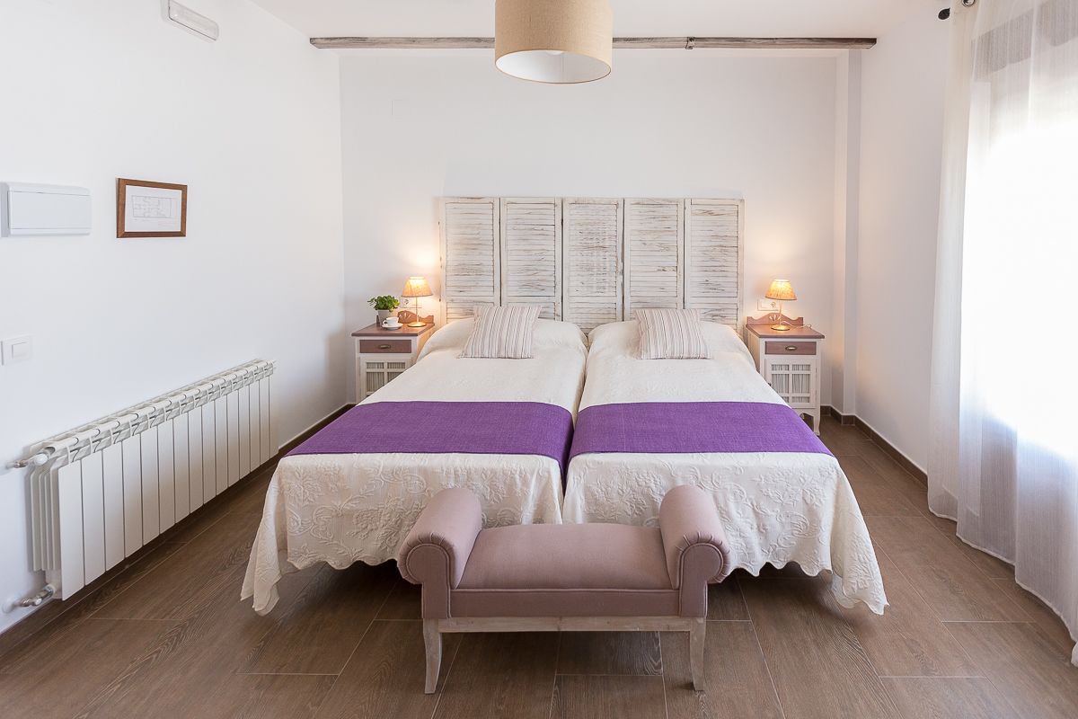 Una cama doble con cabecera de madera, dos mesitas de noche con lámparas, un banco y un radiador.