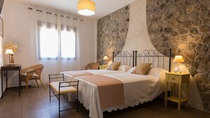 A bedroom with two beds, a stone wall, a window with curtains, and a chair.