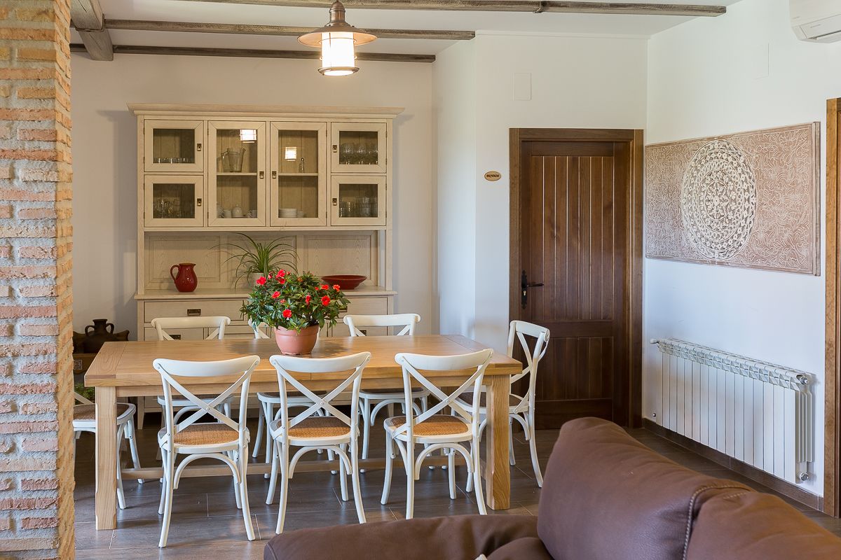 A dining area with a wooden table and white chairs, a potted plant with red flowers, and a brick wall.