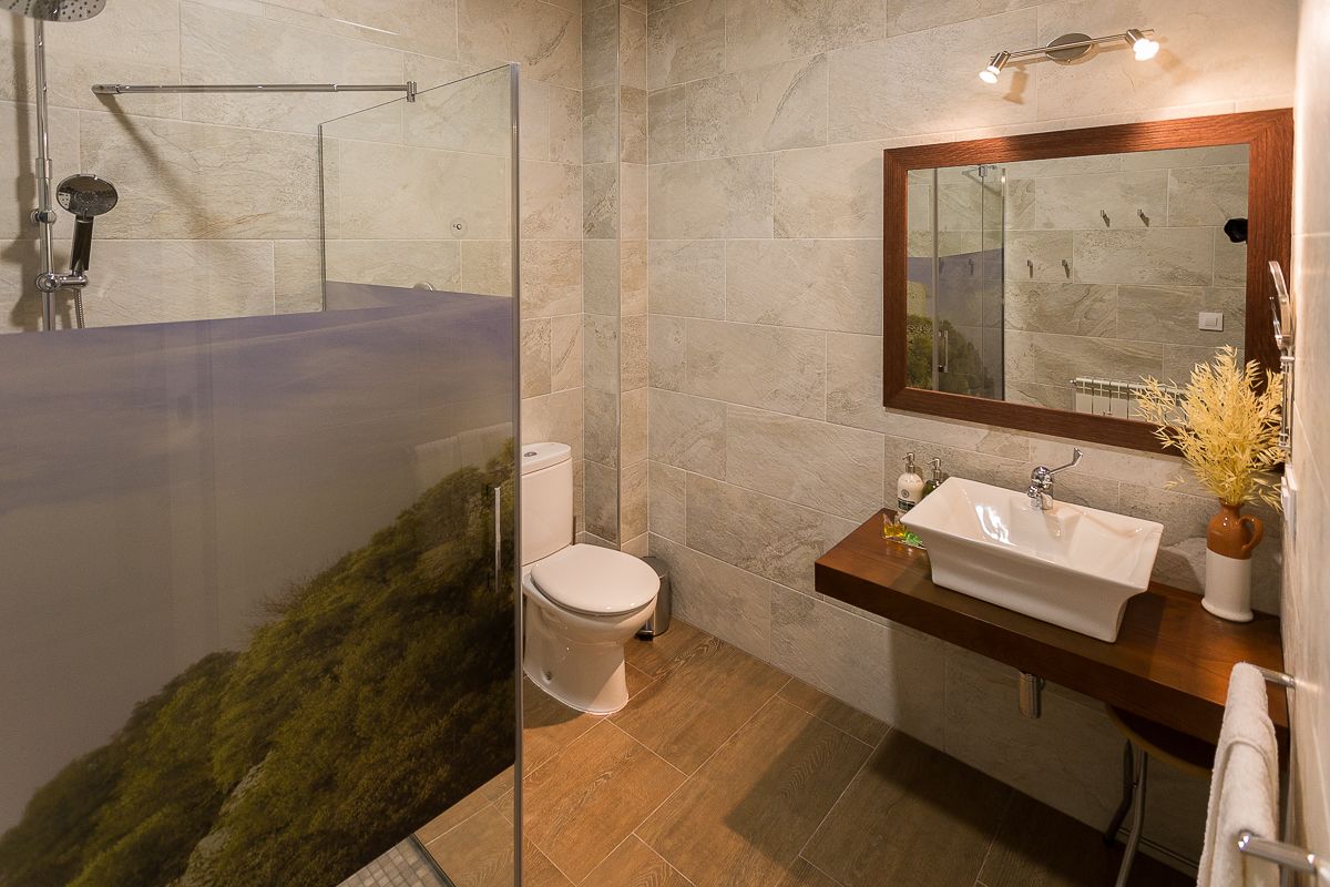 Un baño moderno con ducha de vidrio, inodoro blanco, tocador de madera con lavamanos y espejo sobre el tocador.