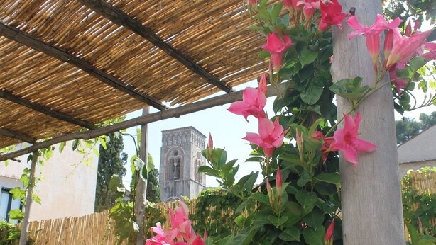 A covered outdoor area with a bamboo roof, pink flowers, and a building with a tower in the background. - Gelsomino Rooms Ravello (Ravello)