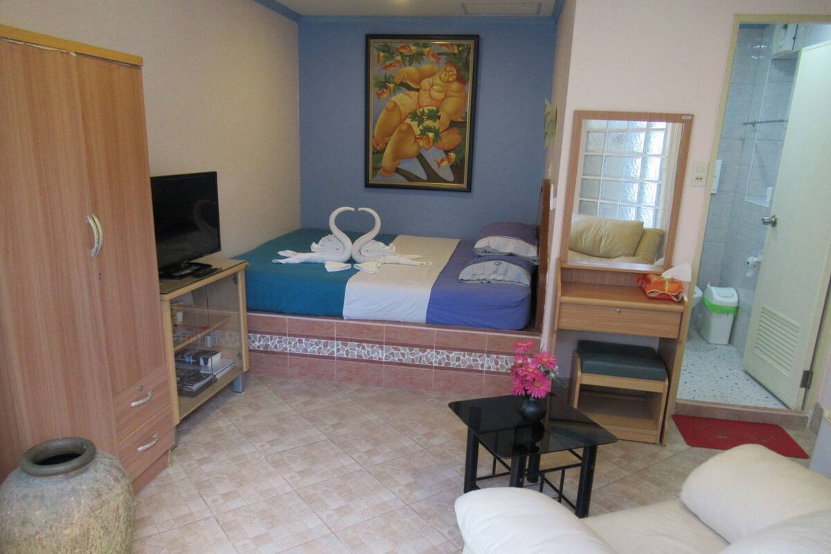 A hotel room with a bed, a television, a desk, a chair, a sofa, a bathroom, and a large vase with flowers.