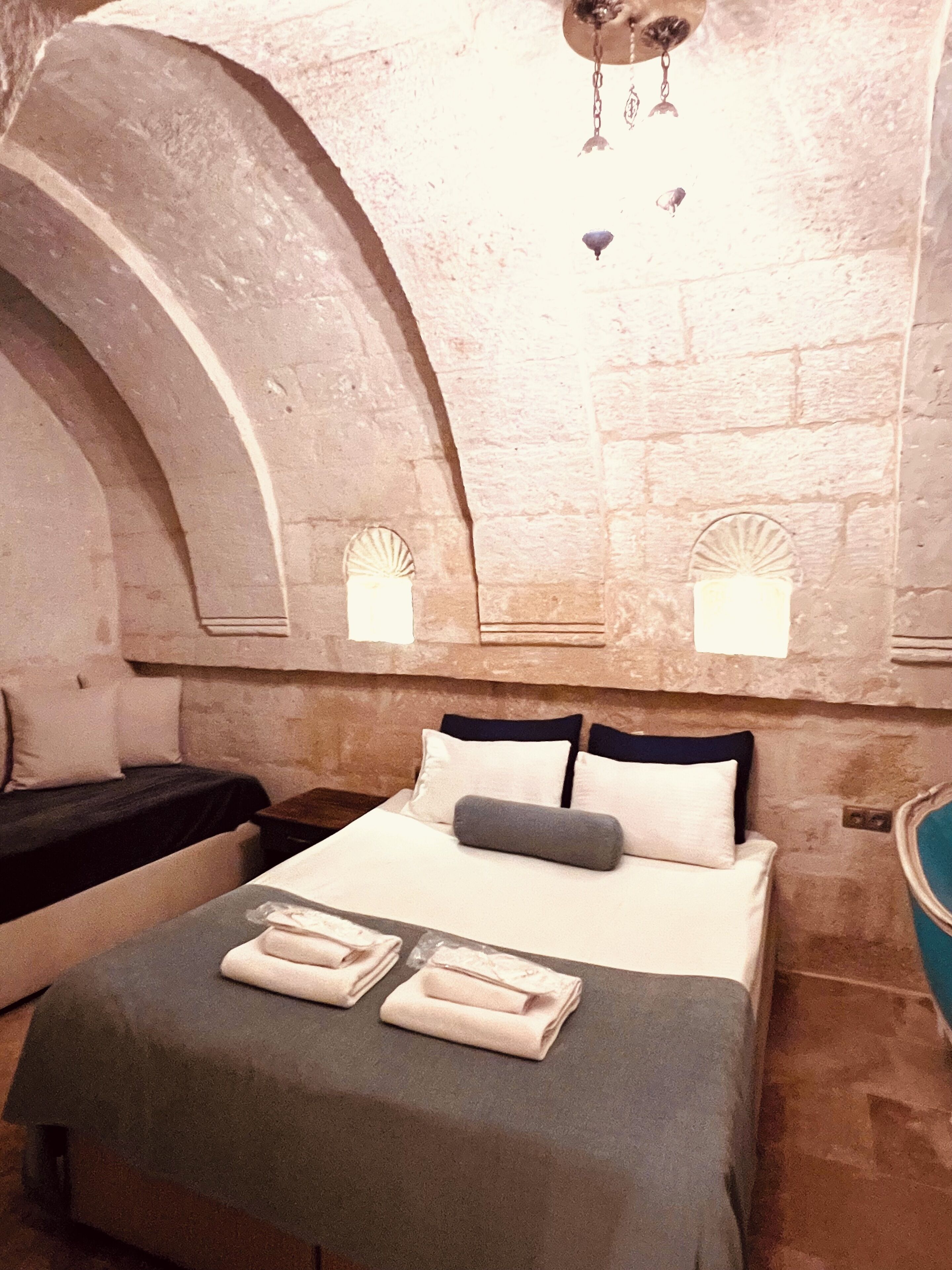 Una habitación de hotel con cama, almohadas, una mesita de noche y una pared de piedra con un ventilador.
