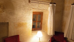 A cozy room with rustic wooden beams, a wooden-framed window, a small round table with a lamp, and two red upholstered chairs. - Roca Cappadocia (Ürgüp)