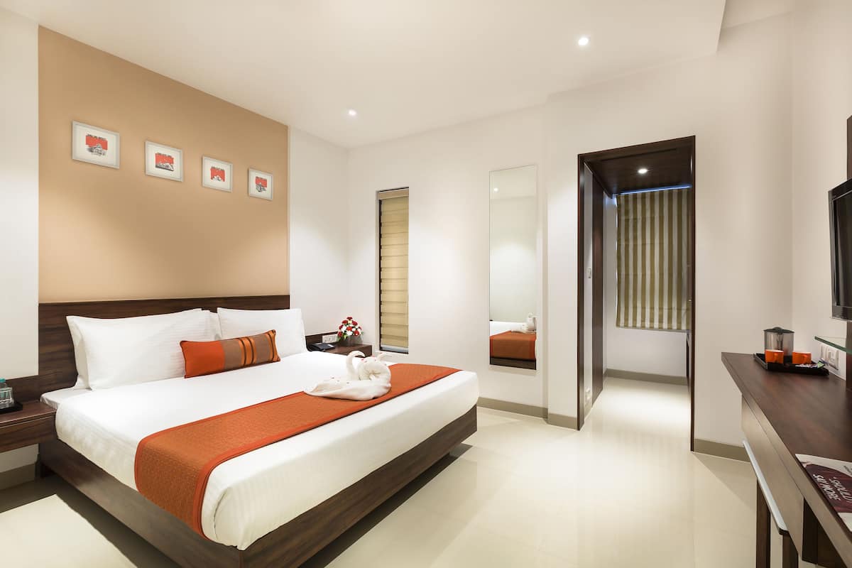 Une chambre d’hôtel moderne dotée d’un grand lit, d’une tête de lit en bois, d’un téléviseur à écran plat, d’un bureau avec une chaise et d’une salle de bain visible à travers une porte ouverte.
