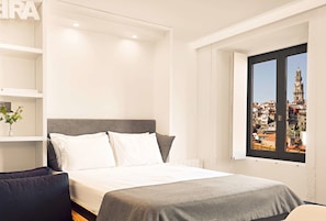 Une chambre d’hôtel moderne, avec un lit, une vue sur le paysage urbain par la fenêtre et une étagère ornée d’une plante.
