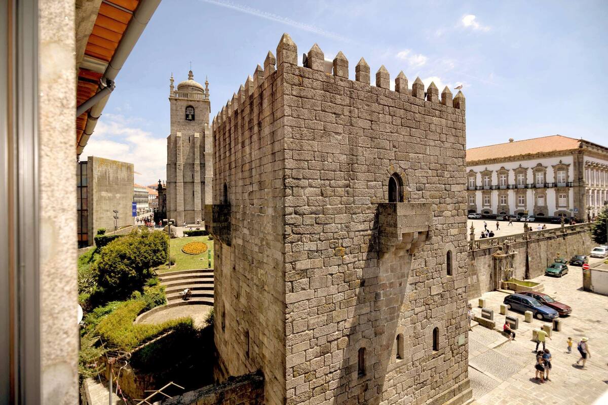 A historic stone tower with battlements, a clock tower in the background, and a paved plaza with pedestrians and vehicles.