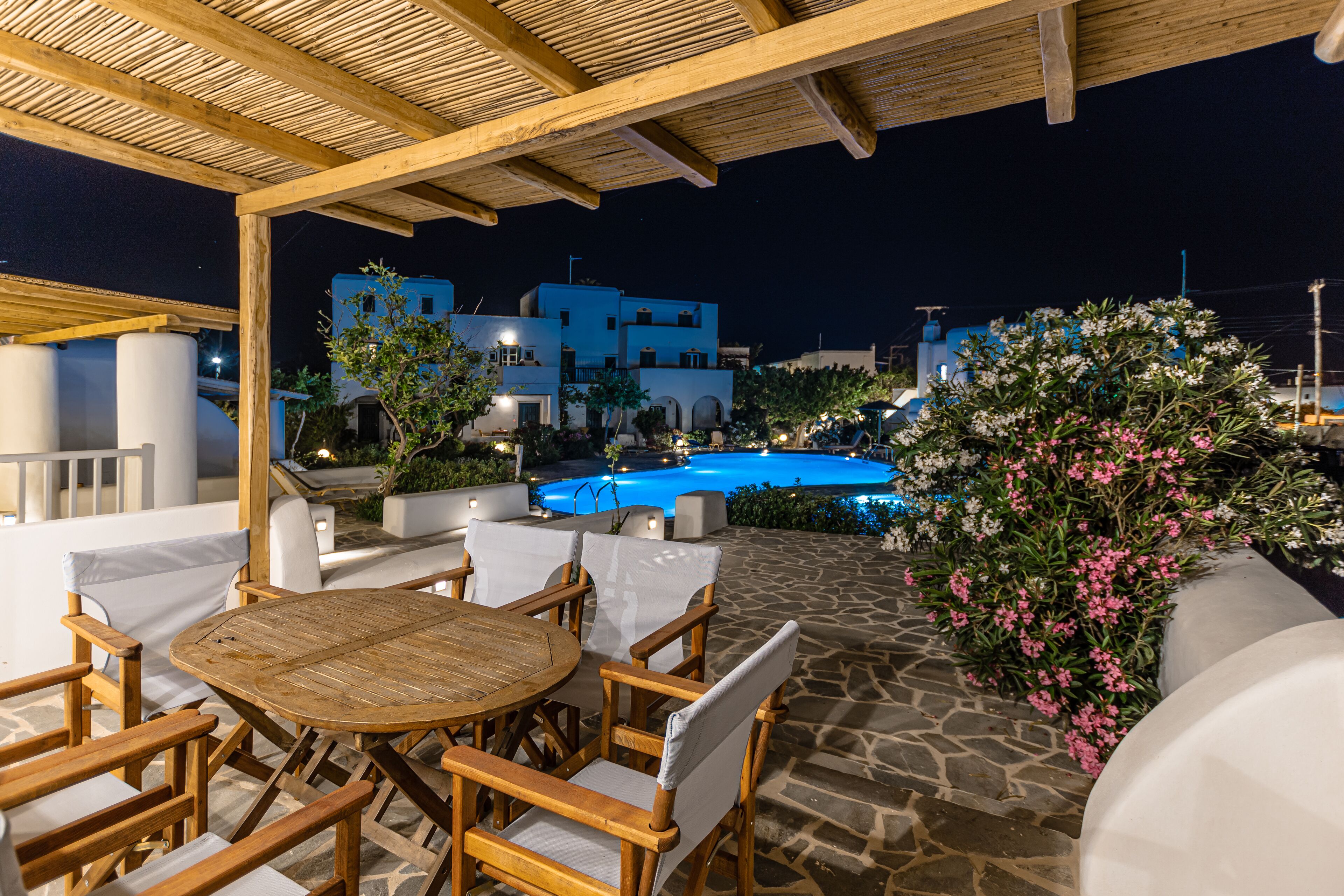 An outdoor patio area with a round wooden table and chairs, a stone floor, and a view of a swimming pool at night.