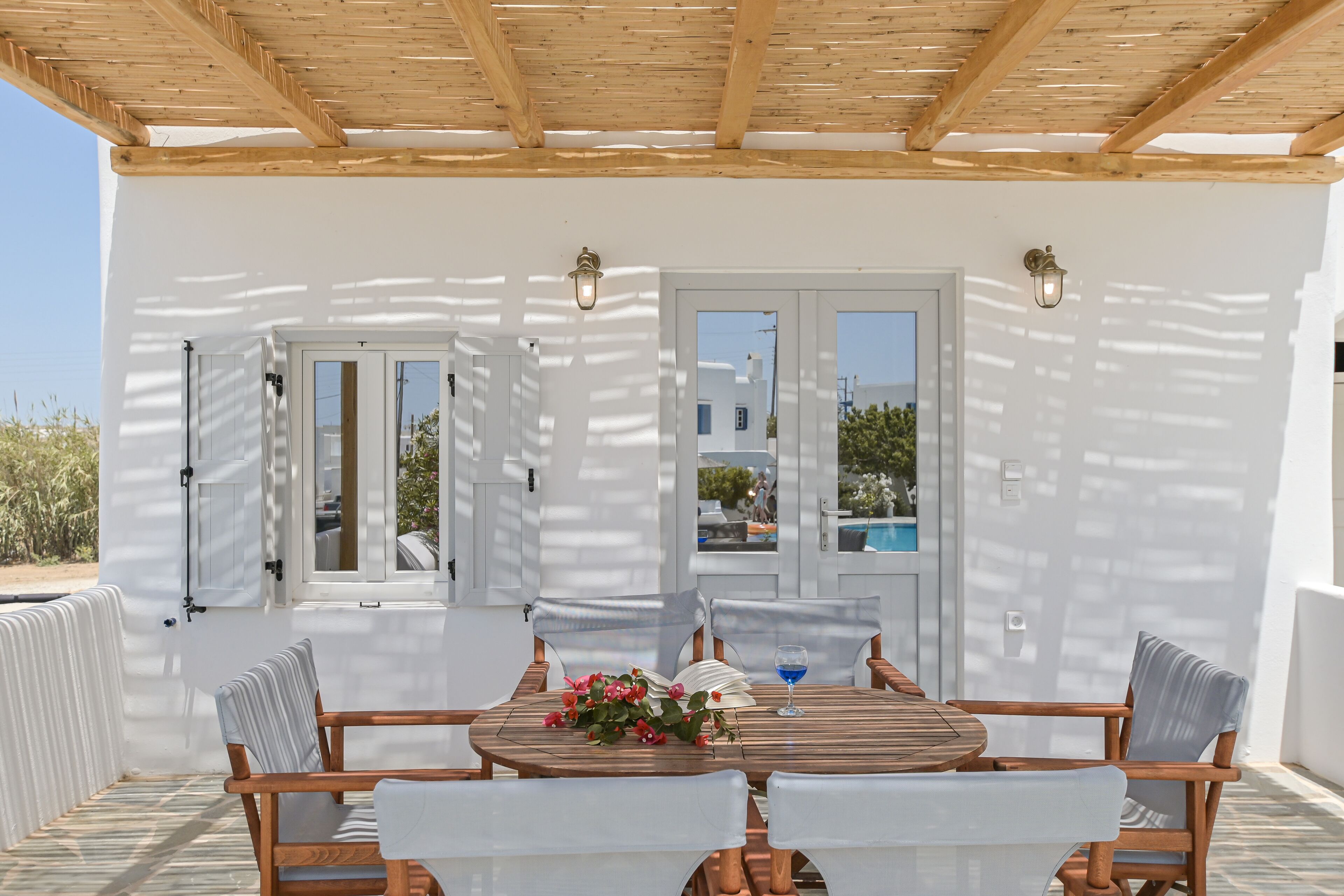 A covered outdoor seating area with a wooden table and chairs, a glass table, and a flower arrangement.
