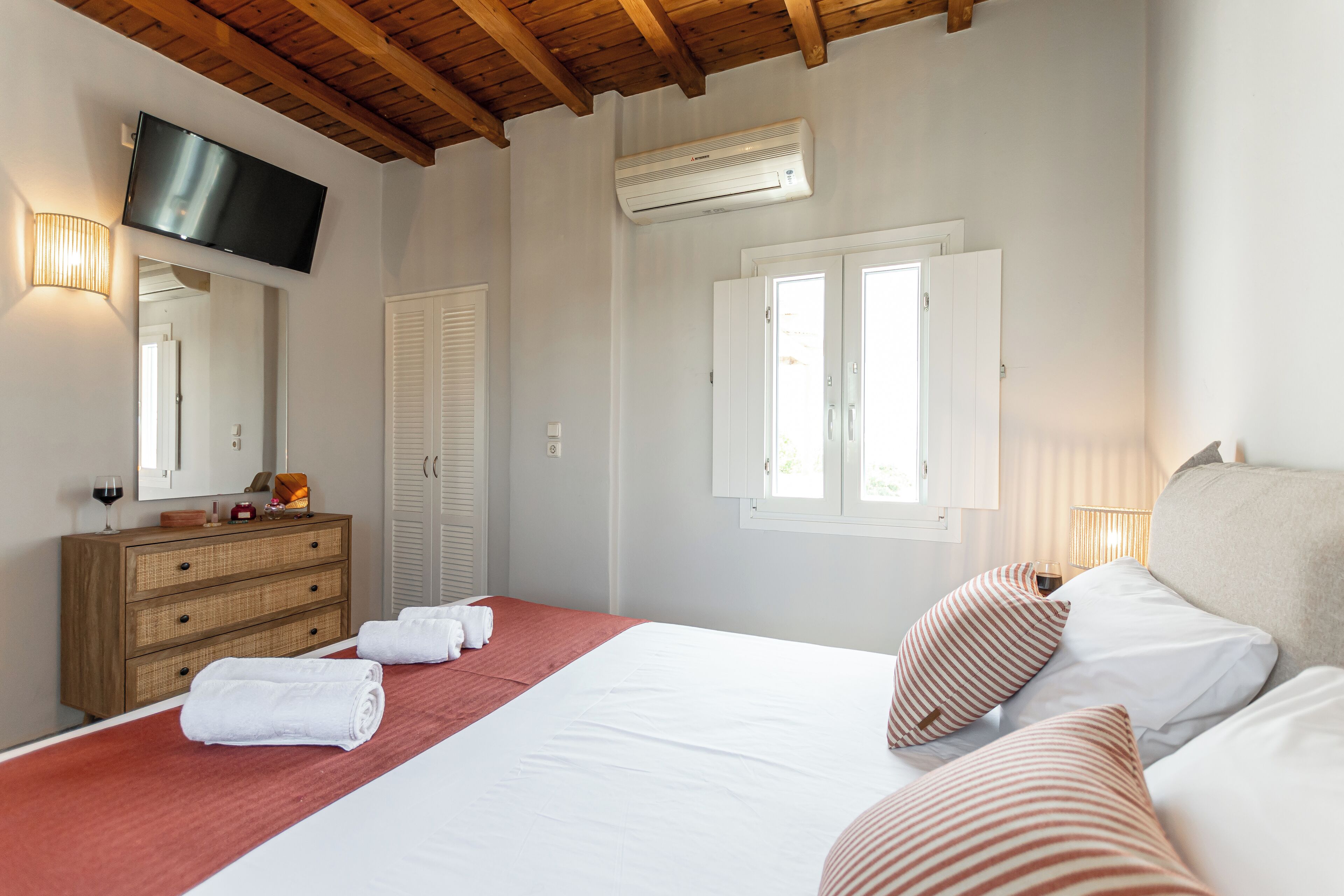 A bedroom with a bed, a TV mounted on the wall, a dresser, and a window with shutters.