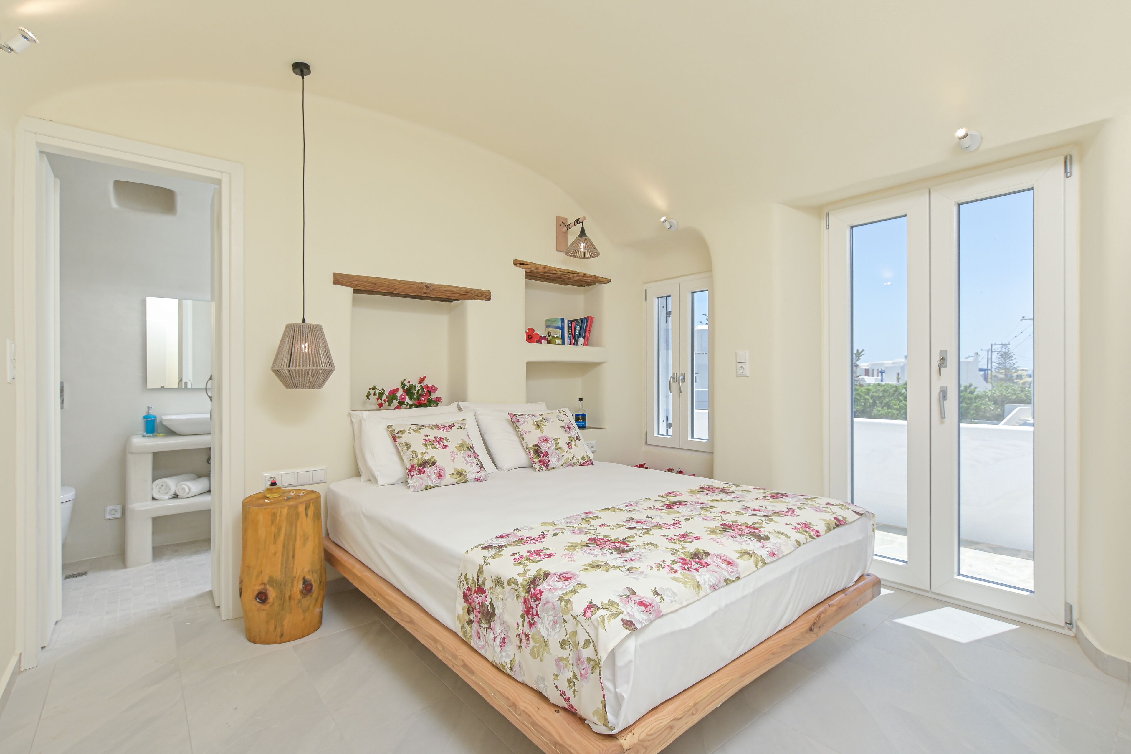 A bedroom with a bed, a wooden stool, a hanging light fixture, a shelf with books, and a view through a door.