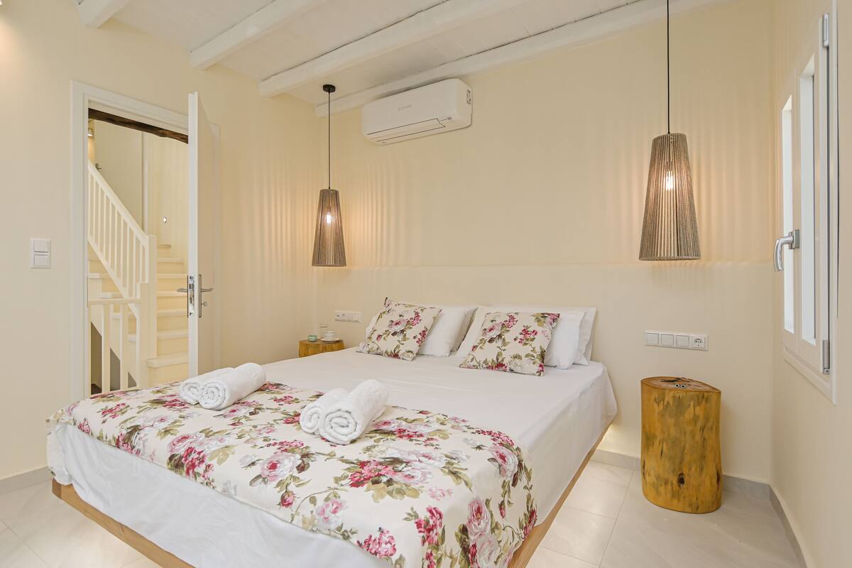 Un dormitorio con una cama, ropa de cama floral, dos mesitas de noche, un equipo de aire acondicionado y un taburete de madera.