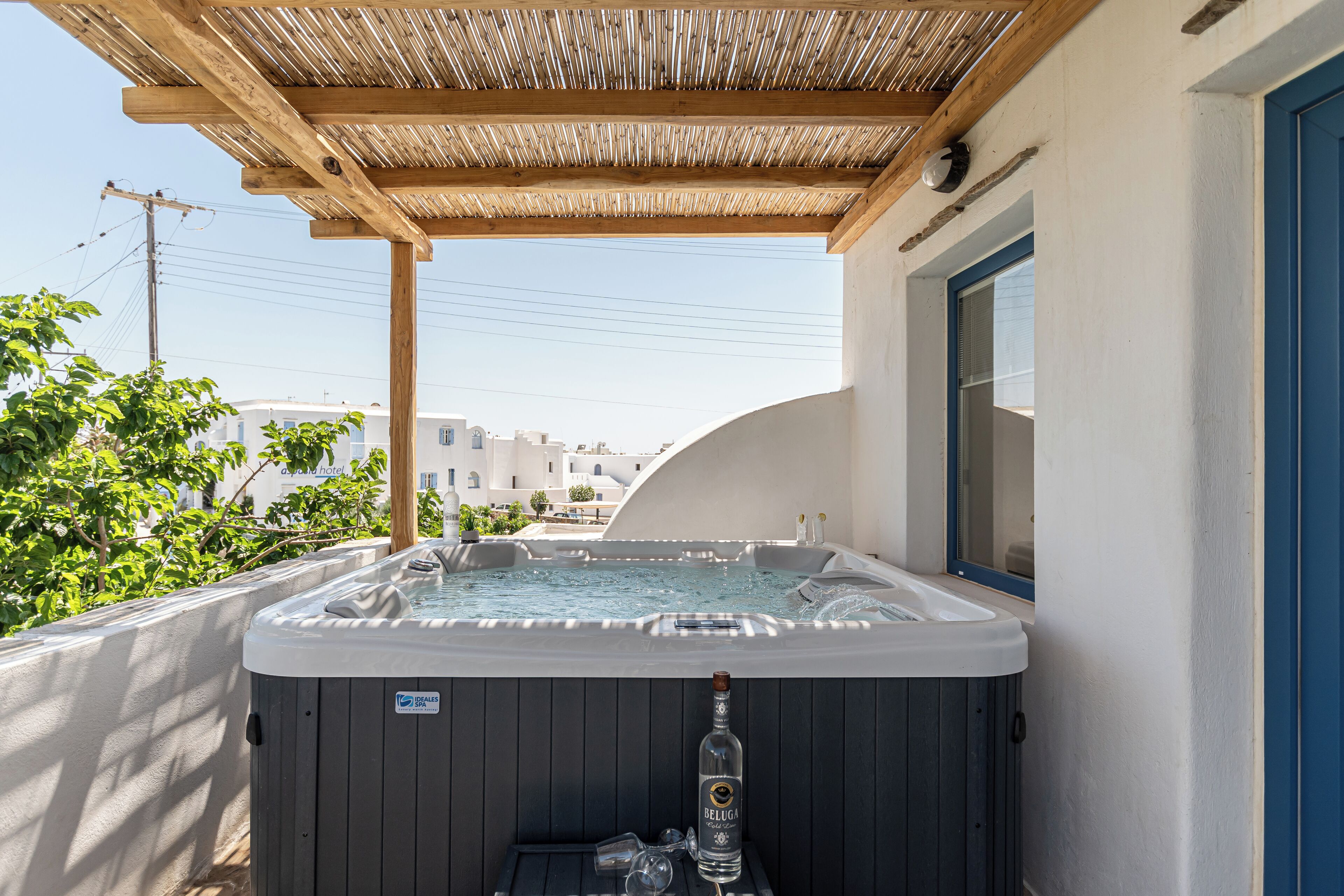 A hot tub with a bottle of Beluga vodka and glasses, under a wooden pergola, with a view of buildings in the background.