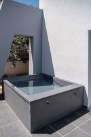 Un moderno jacuzzi exterior con diseño geométrico, frente a una pared blanca y rodeado por un patio empinado.