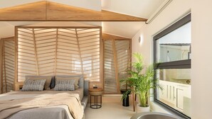 A modern bedroom with a large bed, a wooden headboard, a small side table, a potted plant, and a view of the outside through a window. - Nontas Hotel Apartaments (Chania)