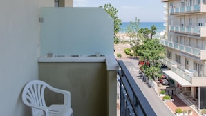 A balcony with a white plastic chair, a view of the sea, and a multi-story building with balconies.