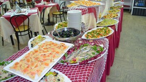 A buffet with various dishes, tables with chairs, and a refrigerator in the background.