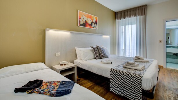 A hotel room with two beds, a bedside table with a lamp, a painting on the wall, and a window with curtains.