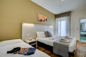 A hotel room with two beds, a bedside table with a lamp, a painting on the wall, and a window with curtains. - Christian Hotel (Riccione)