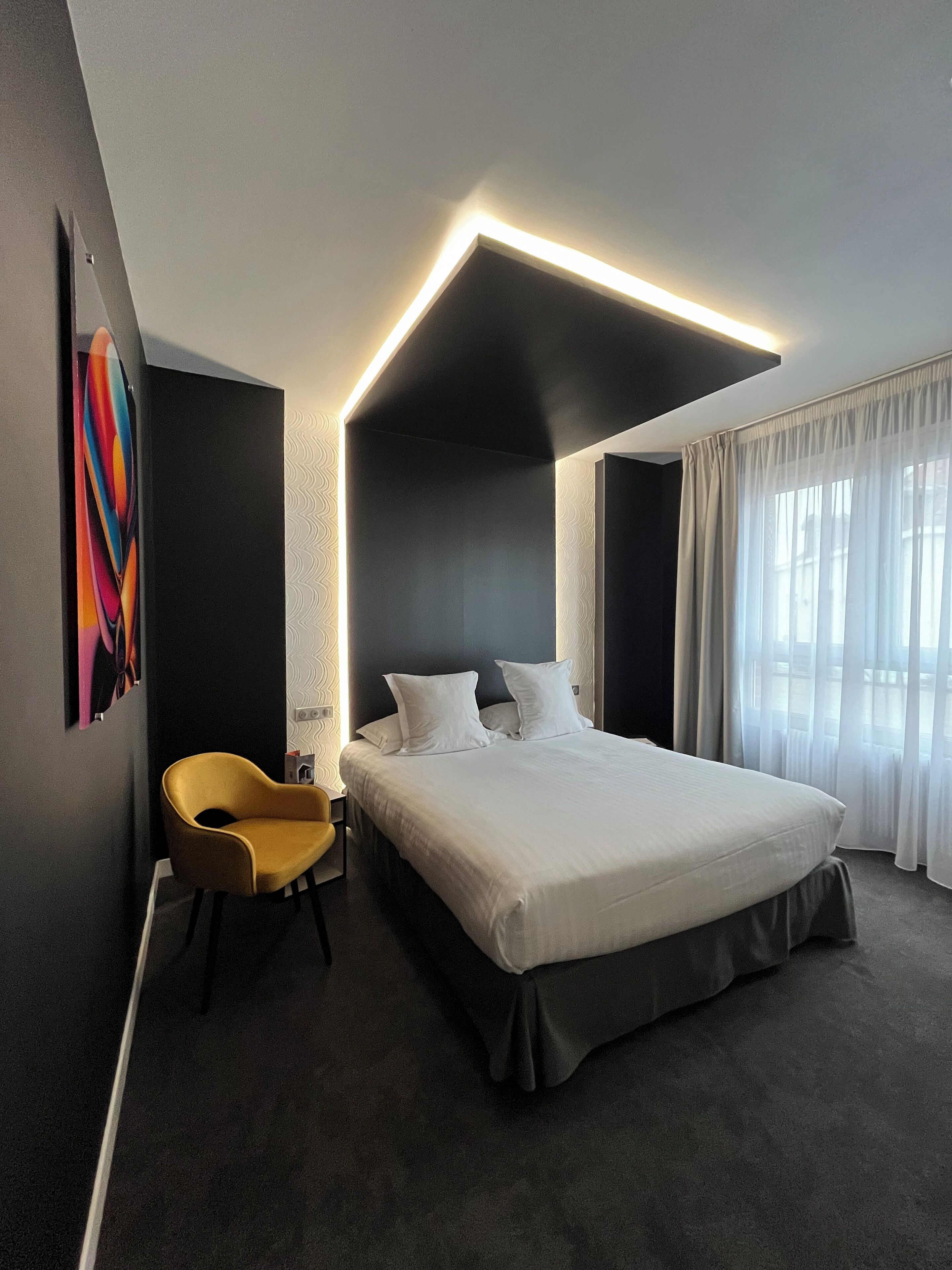 A modern hotel room with a large bed, a yellow chair, and a unique ceiling light fixture.