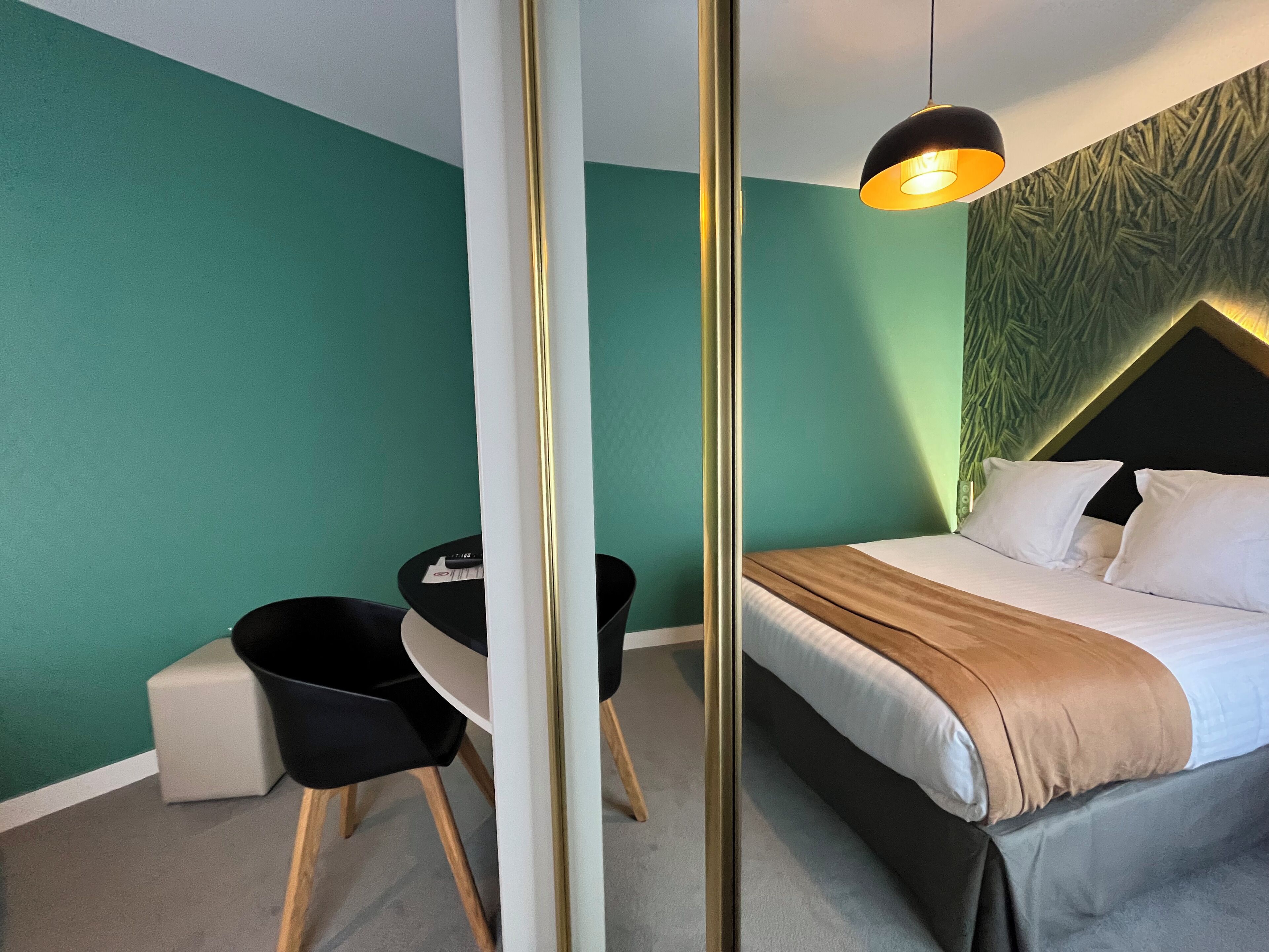 A modern hotel room with teal walls, a bed with white linens, a black desk, and a pendant light.