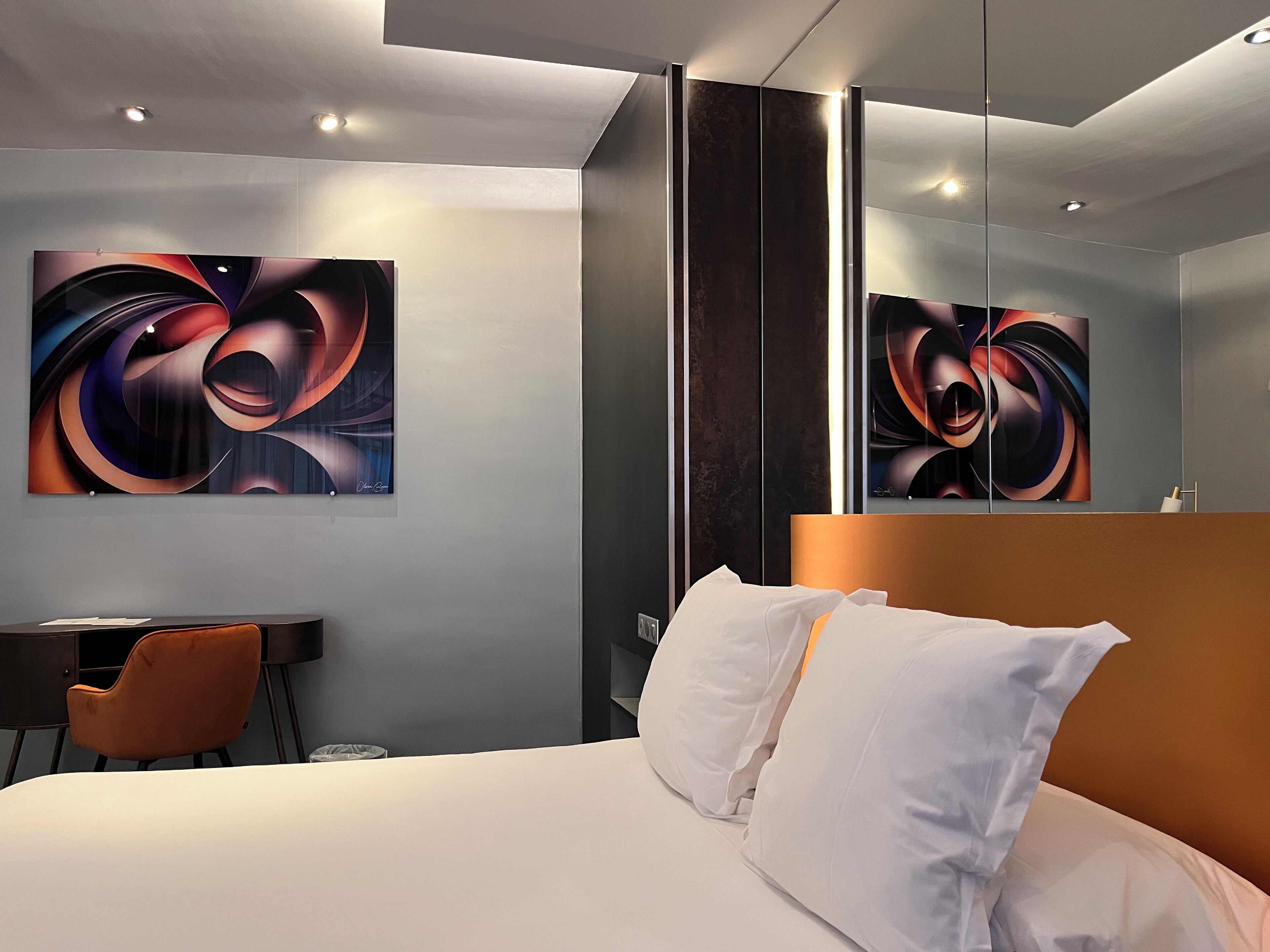 A modern hotel room with a large bed, a desk, and a chair. There are abstract paintings on the walls and a mirror reflecting the room.