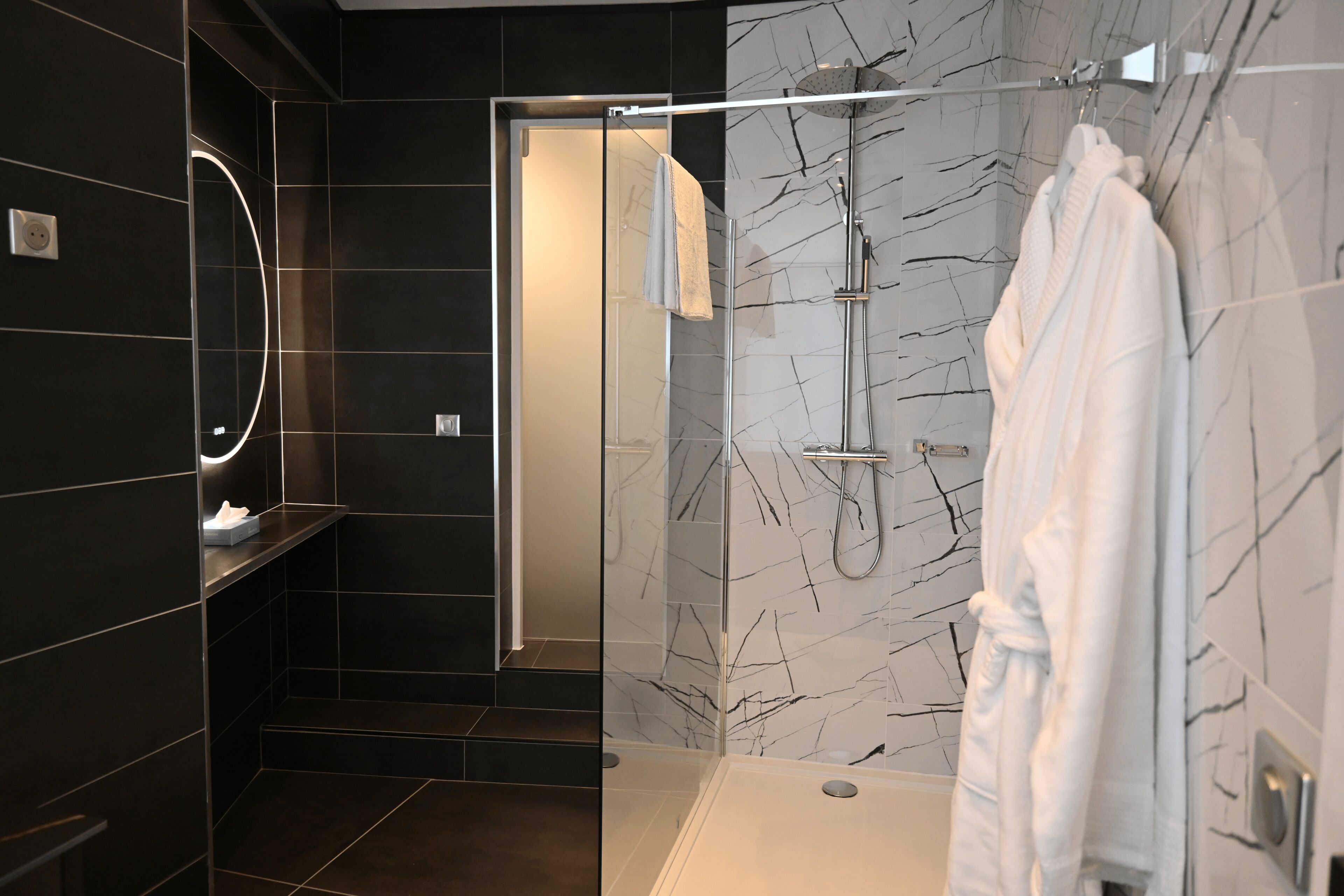 A modern bathroom with dark tiles, a glass shower enclosure, a white robe hanging, and a marble-patterned wall.