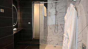 A modern bathroom with dark tiles, a glass shower enclosure, a white robe hanging, and a marble-patterned wall.