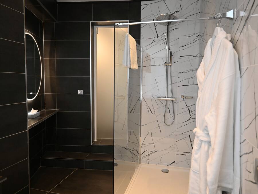 A modern bathroom with dark tiles, a glass shower enclosure, a white robe hanging, and a marble-patterned wall.